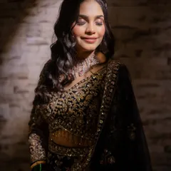 A dramatic and moody portrait. The lighting highlights the intricate embroidery on her dark lehenga and the soft glow of her makeup.