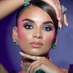 A dramatic beauty shot with vibrant purple eyeshadow and expertly placed highlights. The pose draws attention to the stunning emerald jewelry and flawless manicure.