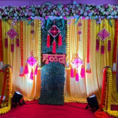 A close-up of a traditional Haldi stage. The decor includes bright yellow drapes, a central green panel, and decorative pink hangings, all brought together with a custom 'हळद' sign to celebrate the occasion.