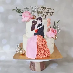 A romantic two-tier anniversary cake featuring a hand-painted couple illustration. The bride's ruffled dress is created with delicate buttercream piping, adding a three-dimensional, artistic touch to the design.