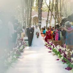 A bride's unforgettable walk down an aisle lined with purple and white flowers. The hanging lights add a magical touch to this outdoor Christian wedding ceremony at Adyar Garden.