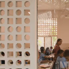 A detail shot looking through a window into a classroom at the rural school. The layers of jaali screens and bamboo roofing create a rich texture and a gentle, diffused light that is ideal for a learning environment.