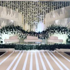 A modern and sophisticated white-themed reception stage. The backdrop uses clean lines, hanging floral installations, and layered panels to create a chic and contemporary ambiance, perfect for a stylish evening celebration.