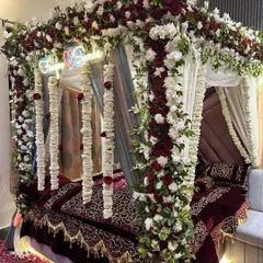 A close-up of a luxurious wedding bed decoration. The four-poster frame is heavily decorated with red and white flowers and hanging floral strings, creating a stunning floral cocoon.