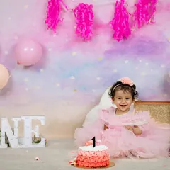 A classic first birthday setup with pink balloons, a beautiful cake, and the word "ONE" in lights. This little girl is all smiles in her lovely pink dress, ready to celebrate.