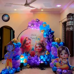 This Krishna theme decoration for a first birthday creates a vibrant and divine atmosphere. The setup includes a large circular backdrop with Yashoda and Krishna, flanked by character cutouts and a rich balloon arch in shades of blue, purple, and gold.