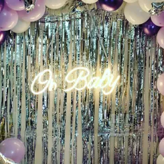An elegant "Oh Baby" baby shower backdrop. We used a silver foil curtain paired with an "Oh Baby" neon sign and framed it with a balloon garland in shades of purple and white for a chic and modern look.