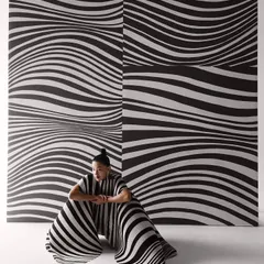 The same concept, with the model seated. The flowing pleats of the dress create a new shape against the rigid graphic background, exploring the contrast between movement and stillness.