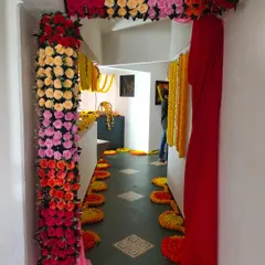 A vibrant entrance decoration for a home event like a haldi ceremony or welcoming the bride. The doorway is framed with colorful artificial flowers and a rich red drape, leading into a hallway decorated with marigolds.