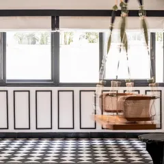 A custom wooden swing with caned details hangs in a sunlit corridor, overlooking the garden. The black and white flooring and paneled walls create a classic colonial feel, perfect for a tropical home.