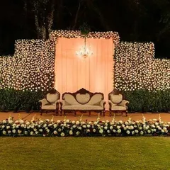 A beautiful Sangeet stage featuring a massive wall of white and red flowers. The vintage-style seating and a central chandelier add a touch of classic elegance to the outdoor lawn setting.