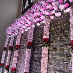 This decor features thick, beautiful flower pillars made of white and red blooms against a rustic backdrop of woven palm leaves. This style is perfect for creating a rich, traditional atmosphere for any pooja or home ritual.