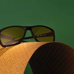 A sophisticated shot of eyewear resting on a curved piece of textured paper. The contrast between the green background and the brown paper creates a rich, earthy color palette.