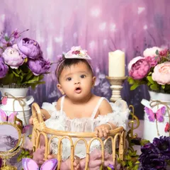 A dreamy, lavender-hued floral session. The baby is dressed in a delicate white lace outfit, sitting in a basket surrounded by purple and pink peonies.