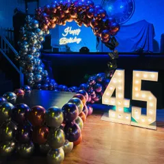 A stylish birthday party backdrop for a 45th birthday. The setup features a circular balloon arch with multi-colored chrome balloons and a 'Happy Birthday' neon sign, complemented by large marquee numbers.