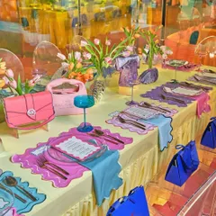 A whimsical and colorful brunch tablescape designed for a couture event. The setting features playful placemats, vibrant glassware, and handbags integrated into the decor, blurring the lines between fashion accessories and table art.