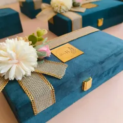 A close-up of the exterior of the premium suede box, adorned with a jute-style ribbon and a floral arrangement. The custom gold acrylic logo tag adds a final touch of brand personalization.