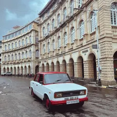 A classic Premier Padmini, with its distinctive red and white paint, parked in front of the beautiful architecture of Horniman Circle.