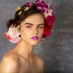 A close-up beauty portrait of a model with fresh flowers woven into her hair and bold purple lipstick. The textured grey background makes the vibrant colors of the makeup and flowers pop.