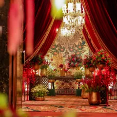 A breathtaking entrance vignette framed by lush red velvet curtains. The space is adorned with a rich tapestry, brass vessels filled with crimson flowers, and a magnificent crystal chandelier, setting a tone of pure opulence.
