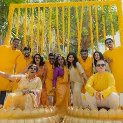 A fun-filled group photo from a Haldi ceremony. The decor is centered around a vibrant yellow theme, with marigold curtains and lotus-shaped seating for the bride and groom.