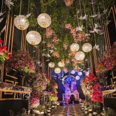 This is an enchanted forest themed entrance passage. The design uses hanging greenery, pink flowers, paper bird ornaments, and woven lanterns to create a whimsical and unique path for guests.