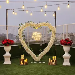 A chic and modern rooftop proposal setup with a stunning city view. The focal point is a large heart-shaped arch of white flowers and greenery, accented with a "Will you Marry Me?" neon sign and surrounded by candles on rustic wood stumps.