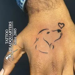 A minimalist line-art tattoo of a dog's face looking up at a tiny heart, placed on the hand. A sweet tribute for any dog lover.