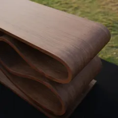 A close-up view of the SWISH bench, highlighting the smooth curves and the rich texture of the wood veneer. The layers of plywood are visible at the edges, revealing the honesty of its construction.