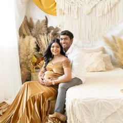 A cozy and romantic couple's portrait in a boho-themed setting. The expecting mother is in a beautiful golden velvet gown, seated with her partner on a decorated bed, creating a warm and intimate memory.
