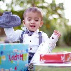 A joyful toddler in a smart bowtie and suspenders smiles while opening birthday presents during an outdoor photoshoot in a park.