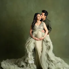 An elegant and romantic portrait of a couple against a textured olive green backdrop. The partner gently kisses the mother-to-be, who is wearing a stunning ruffled gown, showcasing a moment of pure affection.