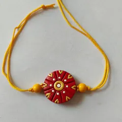 A beautiful terracotta rakhi with a traditional red and gold design on a yellow thread. The intricate dots and patterns give it a festive and auspicious look.