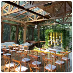 The engagement ceremony space is set within a beautiful glasshouse, with wooden cross-back chairs arranged for guests. The natural light and surrounding greenery perfectly complement the floral decor.