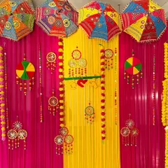 A vibrant Haldi ceremony backdrop. The decor uses colorful drapes in pink and yellow, decorated with traditional Indian umbrellas and floral hangings.