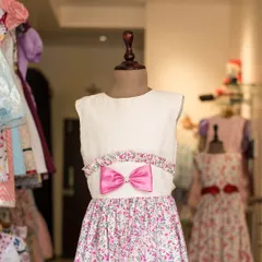 This dress combines a simple white bodice with a vibrant floral skirt. A bright pink bow at the waist adds a fun pop of color, tying the two parts of the dress together.