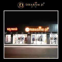 Our storefront at night, with the "Dharam Ji" and "Raymond" signs brightly lit. We are an authorized Raymond dealer, offering their quality fabrics alongside our own bespoke designs.