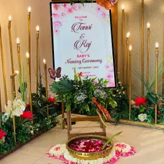 A beautiful welcome sign for a baby's naming ceremony. The decor features a floral and butterfly theme, with elegant candle-like lights and a traditional brass bowl of rose petals.