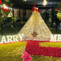 A closer view of the garden proposal setup, highlighting the illuminated 'MARRY ME' letters in front of a decorated teepee tent. The ground is covered with a thick carpet of red rose petals for a luxurious feel.
