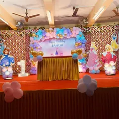 A royal gathering for a first birthday with a Disney Princess theme. The stage features beloved princesses like Ariel, Sofia, and Cinderella, complemented by a pastel balloon arch and a custom backdrop for the little princess, Sanchi.