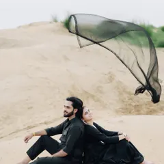 A serene moment in the sand dunes. The couple sits back-to-back while a black dupatta catches the wind, adding a touch of graceful movement to this peaceful portrait.