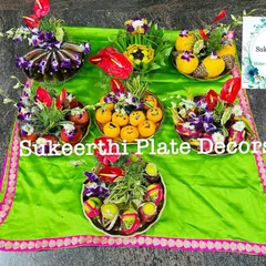An assortment of decorated platters showcasing different styles. You can see arrangements with fresh flowers like anthuriums, fruits like apples and oranges with smiley faces, and beautifully wrapped sweets.