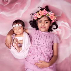 A fun and expressive sibling portrait. The older sister makes a funny face while carefully holding her swaddled newborn sibling against a beautiful floral backdrop.