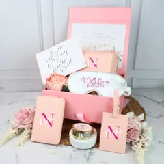 This is my classic pink bridesmaid hamper, filled with personalized travel essentials. It includes a passport holder, luggage tag, and an eye mask, all customized with her initial. It's a practical and beautiful way to ask, "Will you be my bridesmaid?"