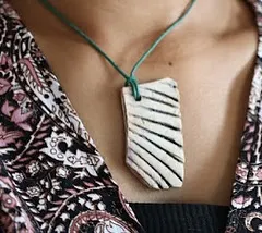 A close-up of a handmade ceramic pendant with a carved line pattern, worn on a simple cord. It's a statement piece that is both modern and earthy.