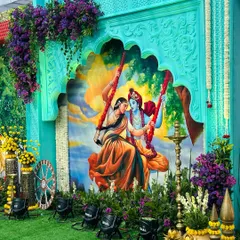 This is a centerpiece from a Vrindavan-themed event, featuring a beautiful custom-painted backdrop of Radha and Krishna on a swing. The setup is framed by intricately carved panels and lush floral arrangements, bringing a divine and artistic feel to the celebration.