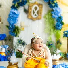 A happy little Krishna sitting on his throne, surrounded by blue flowers and peacock feathers. My setups are designed to be vibrant, authentic, and safe for your baby.