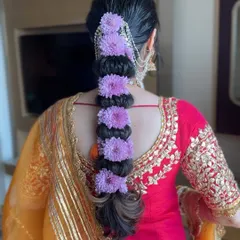 A vibrant and beautiful hairstyle for a Haldi ceremony. This intricate bubble braid is woven with fresh purple flowers, adding a pop of color that complements the festive outfit.