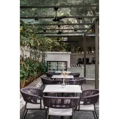 The sun-kissed outdoor seating at Kana Cafe is covered by a glass canopy, with minimal furniture in olive green metal and grey rope. The lime-plastered white walls and concrete flooring enhance the raw, earthy, and natural feel of the space.