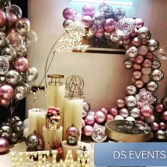 A luxurious party setup featuring a metallic balloon arch in shades of rose gold and silver. The scene is set with elegant white plinths of varying heights, a neon sign, and decorative props like a miniature Ferris wheel.